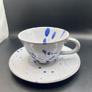 May include: A ceramic teacup and saucer set. The cup and saucer are white with blue splatter paint details. The cup has a handle and sits on a matching saucer. The set is designed for serving tea or coffee.