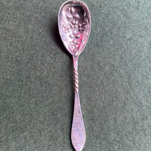 Puede incluir: Cuchara de plata con un diseño detallado de uvas en el cuenco y un mango retorcido. El mango de la cuchara tiene un patrón floral ornamentado en el extremo. La cuchara está sobre un fondo verde oscuro.