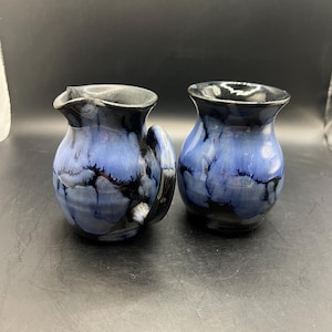 May include: Two ceramic pitchers with a blue and black glaze. One pitcher has a handle and a spout. The other pitcher is a small vase. The pottery has a glossy finish and a unique, handmade appearance.