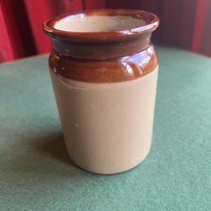 May include: A small, cylindrical ceramic jar. It has a light tan body and a dark brown band around the top, with a dark brown rim. The jar is on a green surface, with a red curtain in the background.