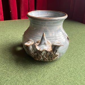May include: A handmade, pale blue ceramic vase with a wide opening and a textured surface. The vase features a relief design of mushrooms and foliage around the base. The rim is a darker brown colour.