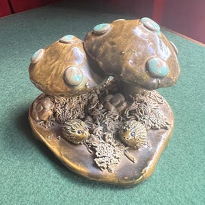 May include: A ceramic mushroom sculpture with two large brown mushrooms and small turquoise accents. The base features small, round creatures and textured details, creating a whimsical, nature-inspired decorative piece.