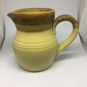 May include: A yellow ceramic pitcher with a brown glaze. The pitcher has a wide mouth and a handle.