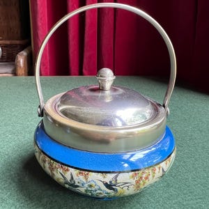 May include: A round ceramic container with a silver-toned metal lid and a matching arched handle. The upper portion is a solid blue, while the lower section features a detailed pattern of colorful hummingbirds, flowers, and foliage on a cream base.