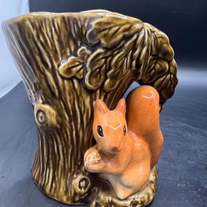 May include: A ceramic planter in the form of a tree trunk, featuring detailed brown bark. An orange squirrel figurine is positioned on the side, holding a nut. The planter has a glossy finish and is designed for decorative use.