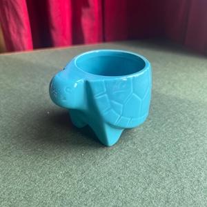 May include: A turquoise ceramic turtle-shaped planter. The planter has a round opening and is designed with a turtle's head, shell, and legs. The shell has a textured pattern. The planter is on a green surface.
