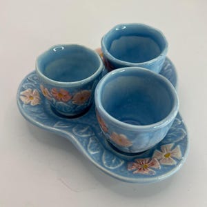 May include: A light blue ceramic tea set with three cups and a matching tray. The cups and tray are decorated with pink and white floral designs. The set is ideal for serving tea or coffee.