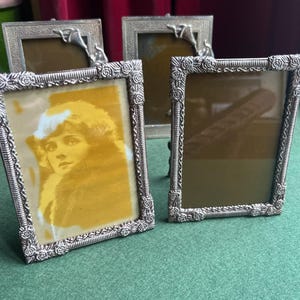 May include: Four antique silver picture frames. The frames have a rectangular shape with ornate floral and scrollwork detailing. One frame contains a sepia-toned portrait. The frames are arranged on a green surface.