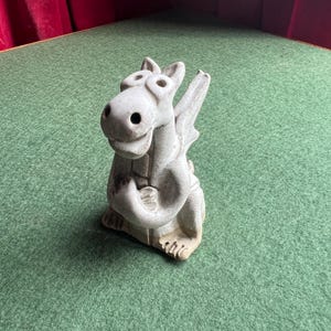 May include: A light grey dragon figurine with intricate detailing. The dragon features wings and a smiling expression, posed in an upright sitting position. It rests on a green surface.