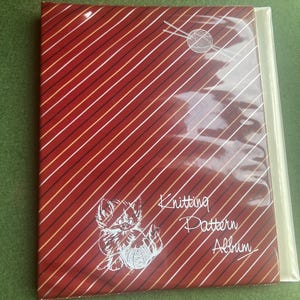 Op de afbeelding: Een rood breipatroonalbum met een diagonaal streepjespatroon in wit, goud en zwart. De omslag toont een witte illustratie van een katje met garen en de woorden "Knitting Pattern Album."