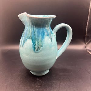 May include: A ceramic jug with a rounded body and a curved handle. The jug is a light turquoise colour with darker blue and green glaze dripping down the sides. The spout is slightly flared.