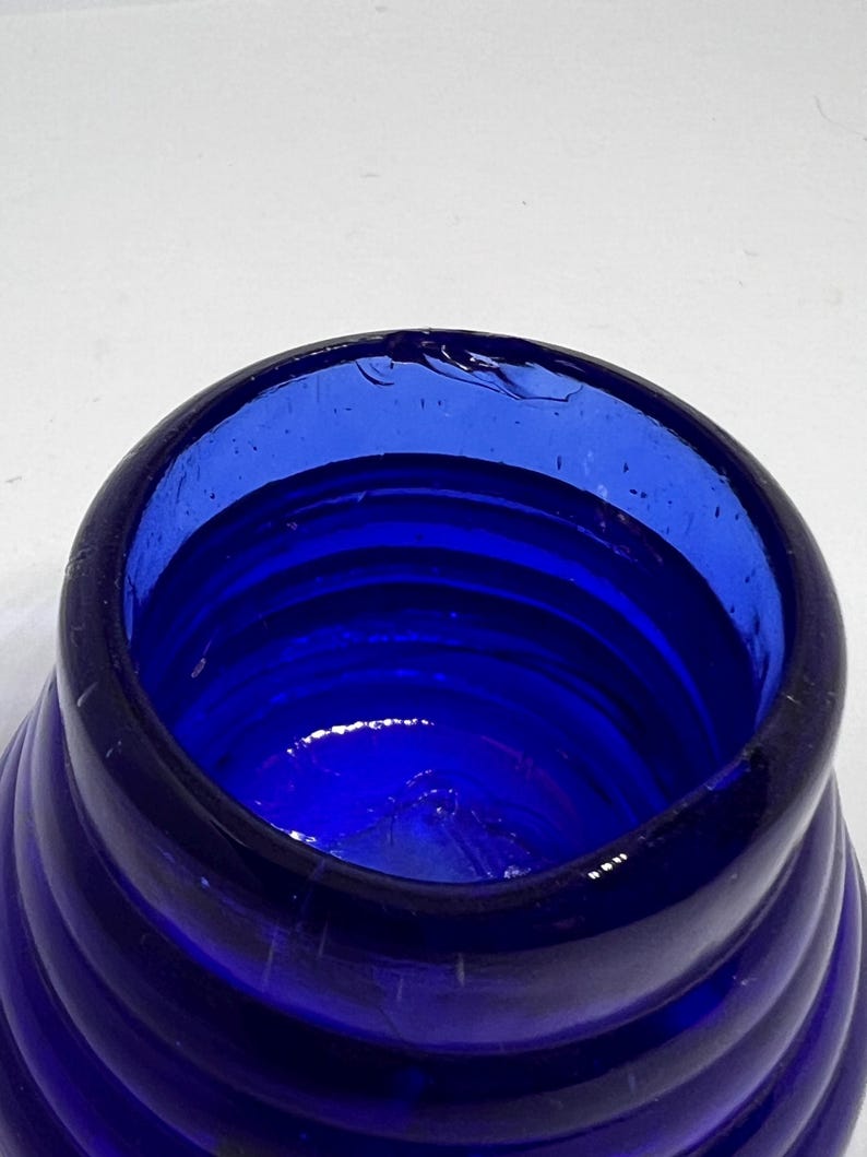 Puede incluir: Primer plano de una botella de vidrio azul cobalto con un dise&ntilde;o en espiral. El borde de la botella tiene una peque&ntilde;a astilla. El vidrio es de un color azul profundo y rico, y el patr&oacute;n en espiral crea un efecto texturizado. Para uso decorativo o funcional.