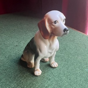 May include: A ceramic beagle dog figurine, seated with its head up. The dog is white with brown and black markings, including brown ears and a black nose. The figurine has a glossy finish and is highly detailed.