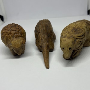 May include: Three small ceramic animal figurines. The figurines are brown and have a rough texture. The animals are a sheep, an echidna, and a dog.
