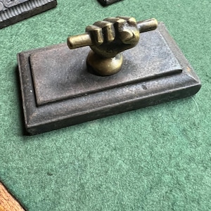 May include: A rectangular, dark metal paperweight with a brass fist handle. The paperweight sits on a green surface, with other similar items visible in the background. The fist handle is detailed and adds a unique touch.