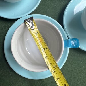 May include: A set of ceramic teacups and saucers. The cups are white inside with a light blue handle. The saucers are light blue. A yellow measuring tape shows the cup is approximately 3 inches in diameter.