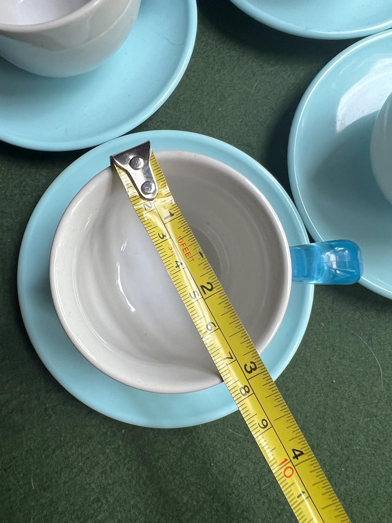 May include: A set of ceramic teacups and saucers. The cups are white inside with a light blue handle. The saucers are light blue. A yellow measuring tape shows the cup is approximately 3 inches in diameter.