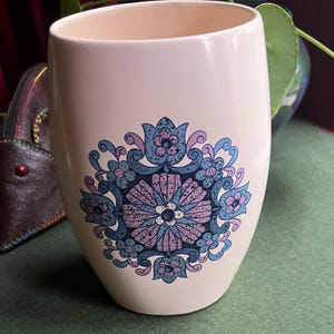 May include: A white ceramic mug with a blue and purple floral design. The design features a central flower with a white center and a ring of smaller flowers around it.