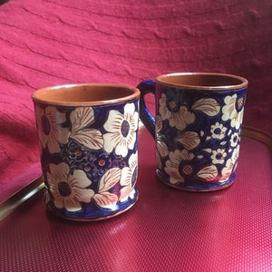 May include: Two ceramic mugs with blue and white floral designs. The mugs have a brown clay rim and a white interior.