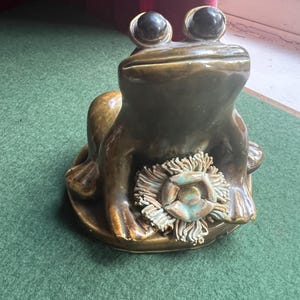 May include: A ceramic frog ornament with large, black eyes and a detailed flower accent. The frog is a mix of brown and gold tones, sitting on a round base. The flower has a turquoise centre and a textured, fringed design.