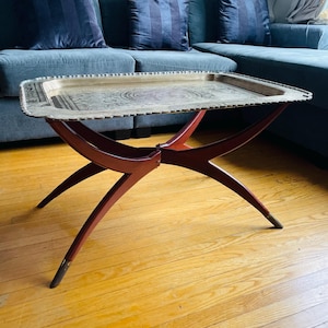 May include: A rectangular brass tray table with a decorative etched design sits on a dark wood base. The base has an X-shaped design with curved legs. The table is placed in front of a blue sofa with matching pillows.