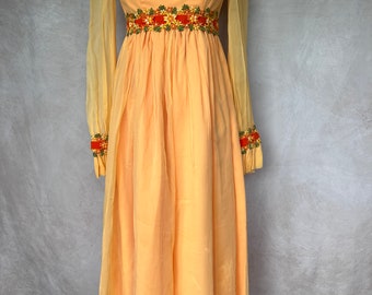 Puede incluir: Un vestido largo y fluido de color amarillo pálido con mangas transparentes. El vestido tiene cuello alto y una banda bordada con flores alrededor de la cintura y los puños.