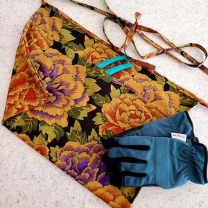 May include: A black gardening apron with a floral print featuring large yellow, orange, and purple flowers. The apron has a long tie and a pocket for tools. A pair of blue gardening gloves are lying on top of the apron.