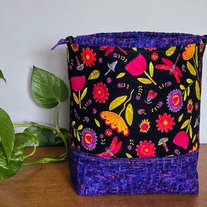 May include: A purple drawstring bag with a black background and a colorful floral pattern. The bag has a purple and black patterned bottom.