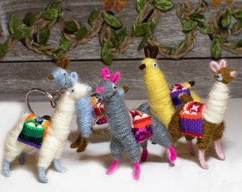 May include: Five colorful llama keychains made of yarn. Each llama has a different colored body and a saddle with a colorful woven pattern. The keychains are on a white surface.
