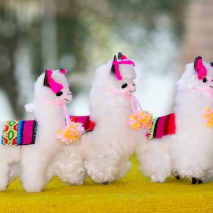 Könnte beinhalten: Drei weiße Alpaka-Plüschfiguren mit rosa und schwarzen Akzenten, bunten Satteldecken und Pompon-Halsketten. Die Alpakas stehen auf einer gelben Oberfläche.
