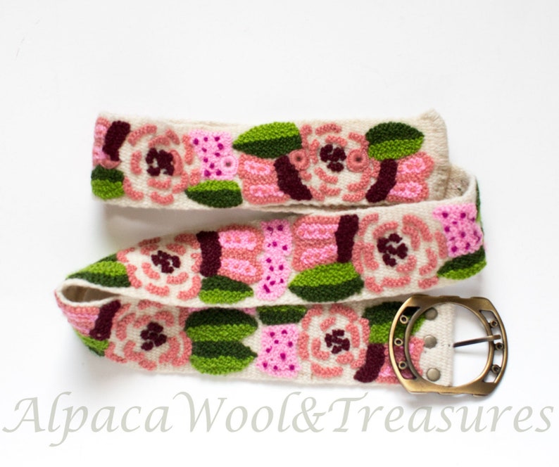 May include: A white alpaca wool belt with a floral design embroidered in pink, green, and burgundy. The belt has a brass buckle.