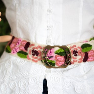 May include: A white embroidered belt with pink and green flowers and a gold metal buckle.