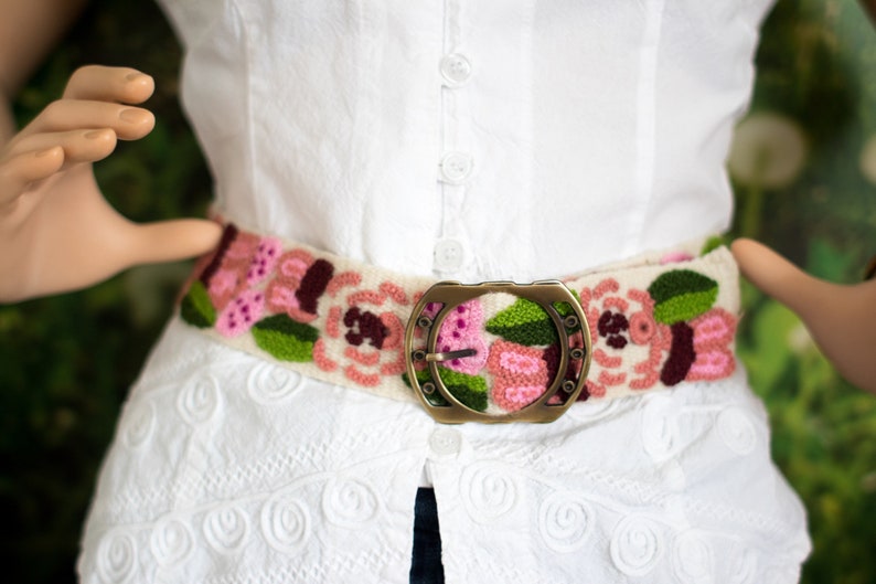 May include: A white embroidered belt with pink and green flowers and a gold metal buckle.