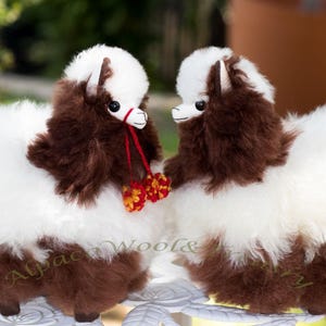 May include: Two fluffy alpaca stuffed animals with white and brown fur. They have small black eyes, white faces, and red and yellow decorative accents. The alpacas are positioned facing each other, suggesting a friendly interaction.