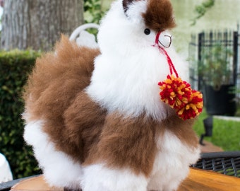 Op de afbeelding: Een bruin en wit alpaca knuffeldier met een rode en gele pompon aan zijn nek.