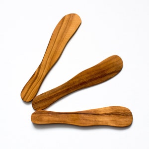 Può includere: Tre coltelli da burro in legno con finitura in legno naturale. I coltelli sono disposti a forma di triangolo su uno sfondo bianco.