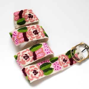 May include: A white fabric belt with a floral design featuring pink, green, and burgundy flowers. The belt has a brass buckle.