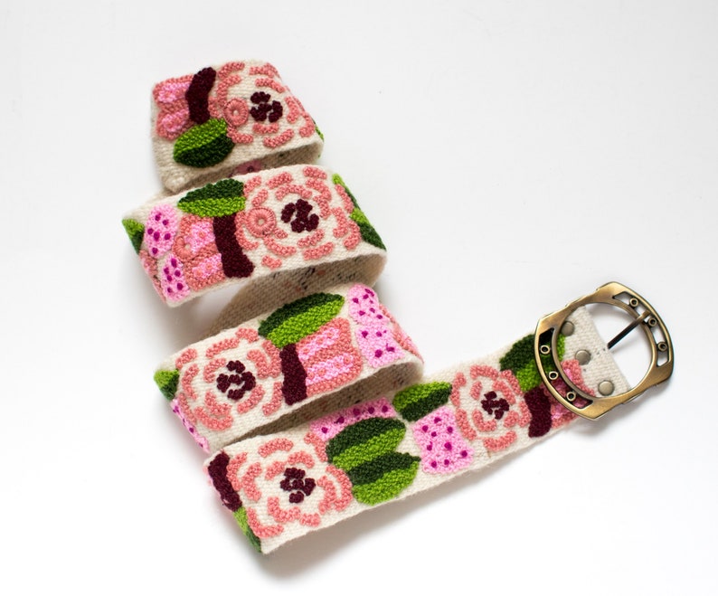 May include: A white fabric belt with a floral design featuring pink, green, and burgundy flowers. The belt has a brass buckle.
