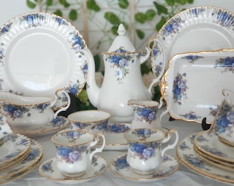 May include: A complete white porcelain tea set with a blue rose pattern and gold trim. The set includes a teapot, cups, saucers, plates, and serving dishes. The edges of the plates have a ridged design.