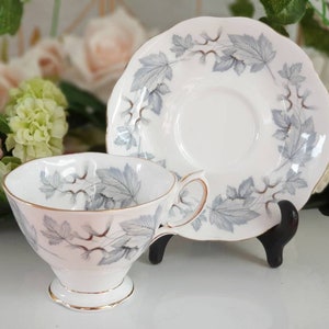 Könnte beinhalten: Eine weiße Teetasse und Untertasse mit einem silbernen Blattdesign. Tasse und Untertasse haben einen goldenen Rand.