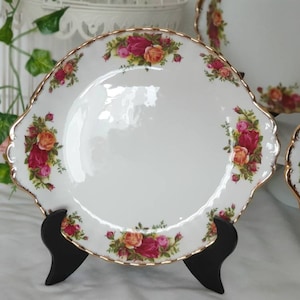 Puede incluir: Un plato ovalado blanco para servir con un borde dorado y un patrón floral con rosas rojas, amarillas y rosas. El plato está sobre un soporte negro.