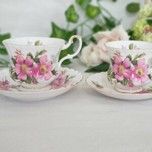 Op de afbeelding: Twee witte theekopjes en schoteltjes met roze rozenbloemmotieven. De kopjes hebben een gouden rand rond de rand.