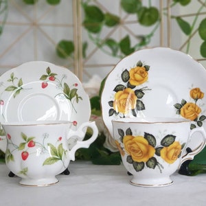 May include: Two white teacups and saucers. One set features a strawberry design with red berries and green leaves. The other set has yellow roses with green leaves. Both sets have gold trim and scalloped edges, perfect for tea parties.