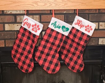 May include: Five red and black buffalo check Christmas stockings with white felt trim and personalised names. The names are "Mom", "Gus", "Aria", "Skip", and "Dad". Each stocking has a green paw print and a green snowflake.