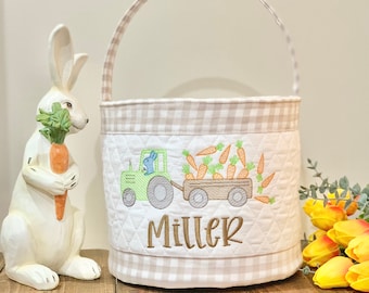May include: A white and tan Easter basket with a quilted design. The basket features an embroidered image of a green tractor pulling a wagon filled with carrots, with the name "Miller" below. A white bunny statue is on the left.