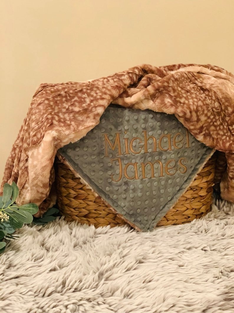 May include: A gray and brown baby blanket with the name "Michael James" embroidered on it. The blanket is folded and placed in a woven basket.