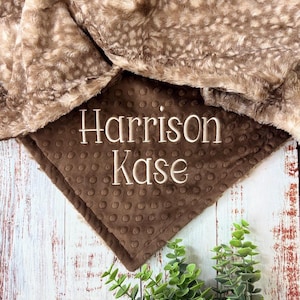 May include: A soft, brown and tan baby blanket with a plush, spotted pattern. The blanket features the embroidered name "Harrison Kase" on a square, textured patch. The blanket is displayed on a rustic, white wooden surface with green foliage.