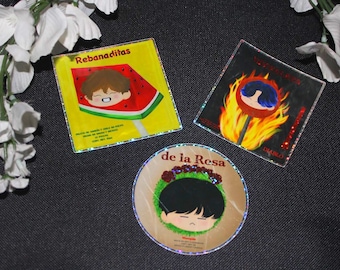 May include: Three holographic stickers featuring cartoon characters with different designs. The first sticker is yellow with a watermelon lollipop and the text "Rebanaditas". The second sticker is black with a red lollipop on fire and the text "No te acerques al Diablo". The third sticker is brown with a wreath of red roses and the text "de la Rosa".