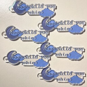 May include: Six clear acrylic charms with a crescent moon and cloud design. Each charm features the text "Moonchild you shine" in a light blue font.