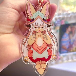 May include: A clear acrylic charm featuring a cartoon character with large ears, glasses, and a detailed outfit in shades of beige, red, and teal. The charm is attached to a gold-colored clasp.